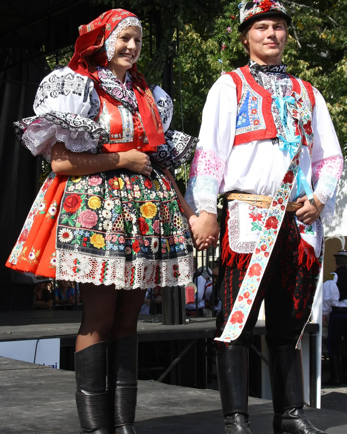 A young couple in festive Moravian Slovácko costume on a stage: a white embroidered blouse and apron with a red headscarf, and a white shirt with a red vest