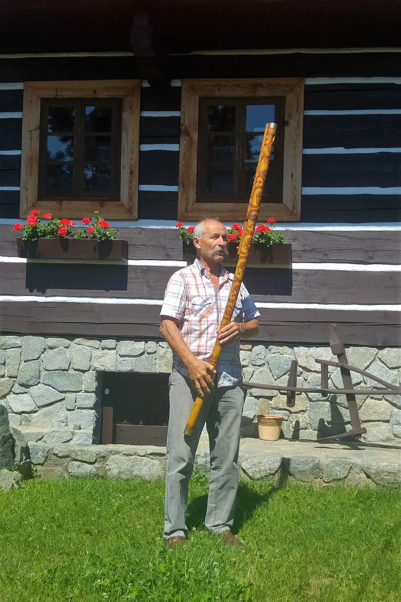 A man standing on a lawn in front of a timber house, holding a fujara, a very long wooden pipe, upright beside him